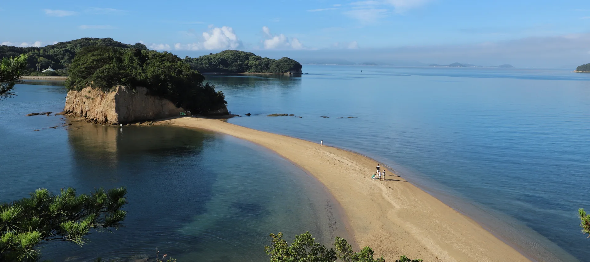 A quiet sandbar curving across blue water toward a wooded island, with a few people walking